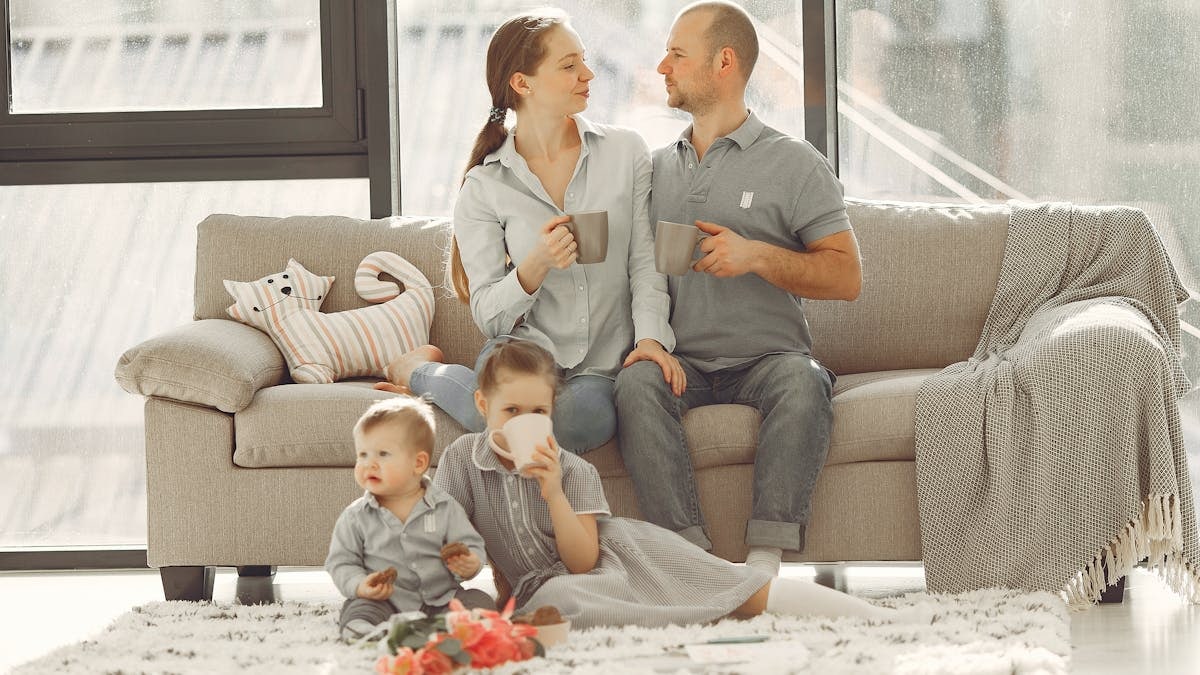Happy family with children spending time at home while parents sitting on couch with cups of coffee and looking at each other when kids resting on floor